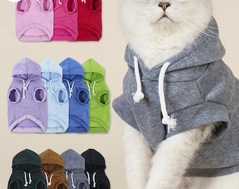 May include: A variety of cat hoodies in various colors, including pink, red, blue, green, and gray. Each hoodie has a hood with a drawstring and a sleeveless design. A gray cat is wearing a gray hoodie.