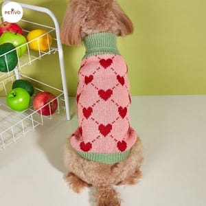 Puede incluir: Un suéter rosa para perros con un patrón de corazones rojos y cuello y puños verdes. El suéter de punto está diseñado para mantener a los perros abrigados y con estilo. Un accesorio para mascotas.