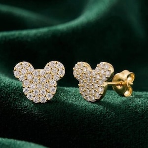May include: A pair of gold-colored stud earrings shaped like a mouse head, encrusted with small, clear gemstones. The earrings are displayed on a dark green, textured surface.