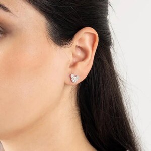 May include: A pair of silver-toned stud earrings in the shape of a mouse head. Each earring is encrusted with small, sparkling crystals. The earrings are shown on an ear.