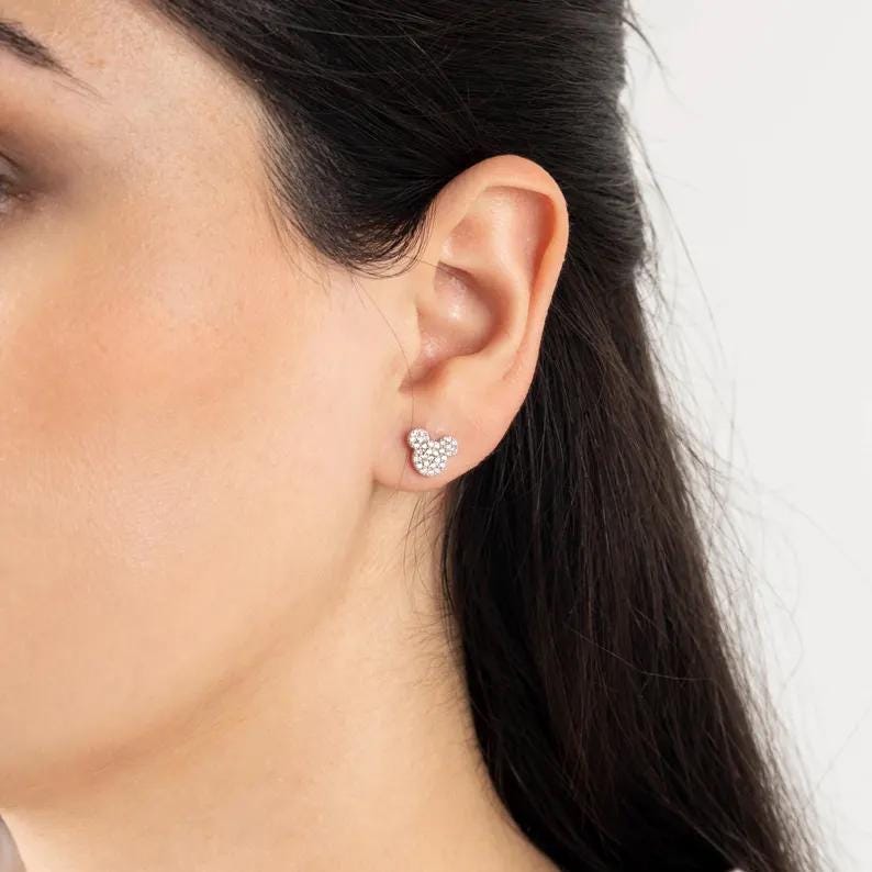 May include: A pair of silver-toned stud earrings in the shape of a mouse head. Each earring is encrusted with small, sparkling crystals. The earrings are shown on an ear.