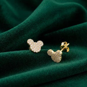 May include: A pair of gold-colored stud earrings in the shape of a mouse head. The earrings are covered in small, sparkling stones, and are set against a dark green velvet background.