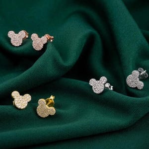 May include: Several pairs of stud earrings in the shape of a mouse head are displayed on a dark green fabric. The earrings are in gold, silver, and rose gold tones, all encrusted with small, sparkling stones.