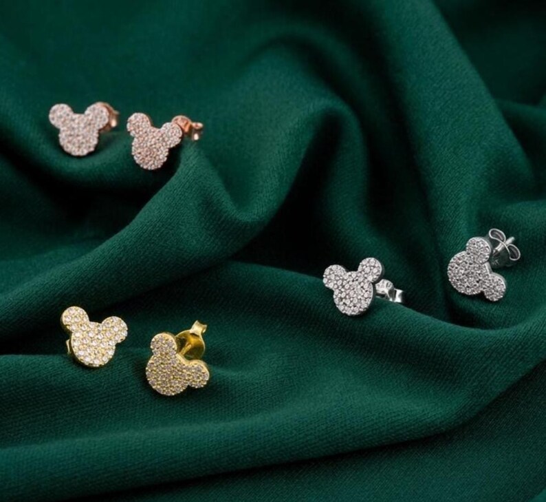 May include: Several pairs of stud earrings in the shape of a mouse head are displayed on a dark green fabric. The earrings are in gold, silver, and rose gold tones, all encrusted with small, sparkling stones.