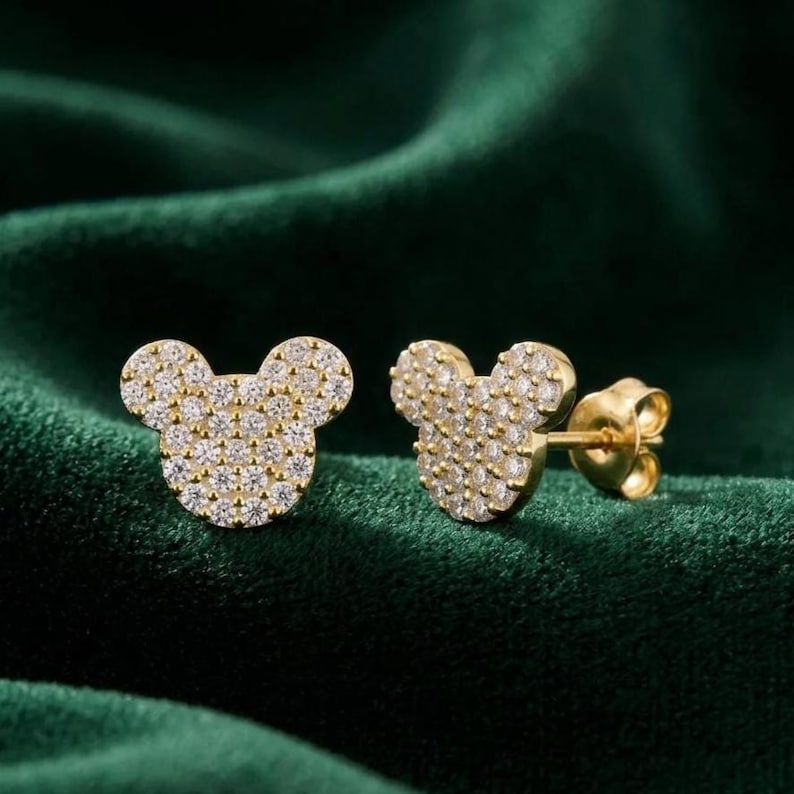 May include: A pair of gold-colored stud earrings shaped like a mouse head, encrusted with small, clear gemstones. The earrings are displayed on a dark green, textured surface.