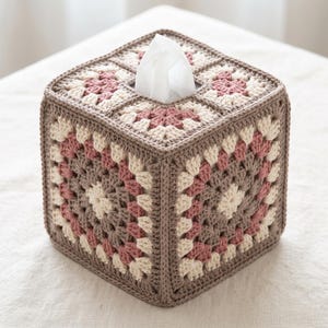 Puede incluir: Una funda cuadrada para caja de pañuelos tejida a ganchillo con un patrón de cuadrados de la abuela. El diseño presenta vueltas alternas de hilo rosa empolvado, crema y topo. Un pañuelo blanco sobresale por la abertura central superior.