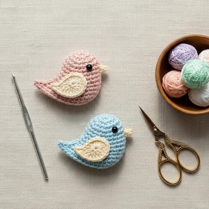 Pode incluir: Dois ornamentos de pássaros de crochê, um rosa e outro azul, com asas e bicos creme. Também estão presentes uma agulha de crochê, tesouras e uma tigela de madeira com novelos de lã coloridos. Os pássaros têm cerca de 10 cm de comprimento.