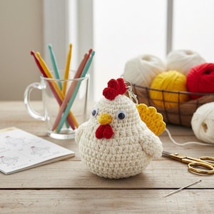 Może przedstawiać: Ręcznie robiony brelok do kluczy z szydełkowym kurczakiem z czerwonym grzebieniem i koralami, żółtym dziobem i ruchomymi oczami. Kurczak jest kremowy z żółtym skrzydłem. Szydełka w szklanym kubku i kulki przędzy w tle.
