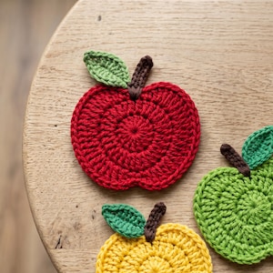 Può includere: Tre sottobicchieri all'uncinetto a forma di mela nei colori rosso, verde e giallo. Ogni sottobicchiere presenta un corpo circolare con un gambo marrone e una singola foglia verde attaccata alla parte superiore. La trama del filo è visibile.