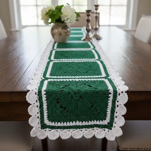 May include: A handmade green and white crochet table runner. The runner features a series of green squares with a white border and a scalloped white edge. The runner is displayed on a dark wooden table, with a vase of white flowers in the background.