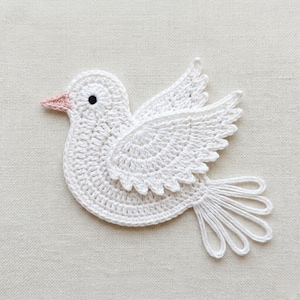 May include: A white crochet dove with a pink beak and a black eye. The dove has detailed wings and a tail, all crafted from white yarn. The handmade bird is set against a neutral background.