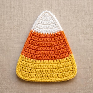 May include: A triangular crochet piece shaped like a candy corn, featuring a white top section, a wide orange middle band, and a yellow base. The item is constructed with visible yarn stitches in a consistent pattern throughout the entire object.