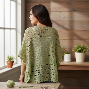 May include: A light green, open-weave crochet shawl with a scalloped edge. The shawl is worn over a white top. The image is taken indoors, with a wooden background and a window to the left. A ball of yarn and crochet hook are on the table.