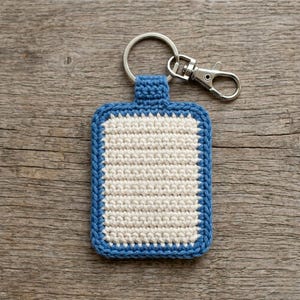 Puede incluir: Un llavero rectangular de ganchillo con un panel central de color crema bordeado por un marco de hilo azul grueso. La pieza está unida a una anilla de metal plateado y a un cierre de mosquetón para sujetar las llaves de forma segura.
