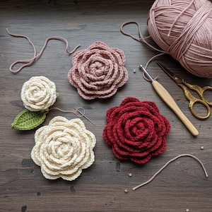 May include: A collection of handmade crochet flowers in various colors, including cream, dusty rose, and deep red. A green crochet leaf and a ball of yarn are also present, along with a crochet hook and gold scissors, suggesting a crafting theme.