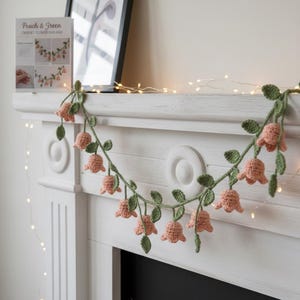 May include: A crocheted flower garland with peach-colored bell-shaped flowers and green leaves. The garland is draped across a white mantelpiece. A card with the text "Peach & Green CROCHET FLOWER GARLAND" is visible.
