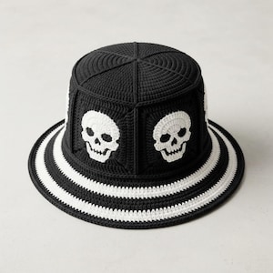 Peut inclure: Un chapeau bob en crochet noir orné de motifs de crânes blancs intégrés sur les panneaux latéraux. Le chapeau présente un sommet plat et un large bord décoré de rayures horizontales noires et blanches alternées sur toute la circonférence.