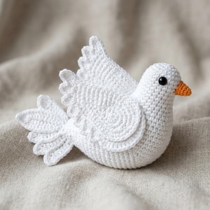 以下が含まれることがあります： 白いかぎ針編みの鳥の置物。翼と尾には質感のあるステッチ模様が施され、小さなオレンジ色のくちばしと黒いビーズの目が一つ付いています。鳥は翼を少し持ち上げて広げた状態で止まっている姿で表現されています。
