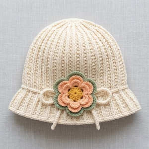May include: Cream-colored crochet hat with a ribbed design and a small brim. The hat is adorned with a crocheted flower in shades of peach, green, and yellow. The flower is centered on the front of the hat, adding a decorative touch.