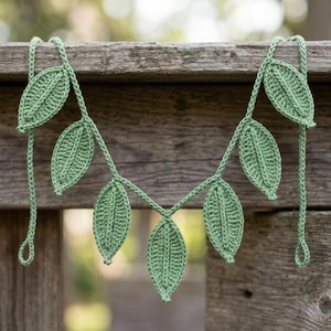 May include: A sage green crochet garland featuring six individual leaf-shaped motifs attached to a central braided cord. Each leaf is crafted with a visible central vein and textured stitch pattern, with small loops at both ends of the cord.