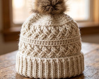 May include: A cream-colored crocheted beanie featuring a thick ribbed brim and a textured cable knit pattern throughout the body. The hat is topped with a large, fluffy brown faux fur pom-pom, creating a classic winter accessory design.