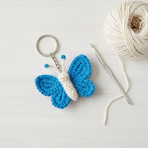 May include: A blue and white crocheted butterfly keyring. The butterfly features blue wings and a white body, attached to a silver key ring. A ball of cream-coloured yarn and a crochet hook are also in the image.