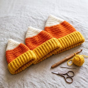 May include: Three crocheted hats in a candy corn pattern with white tips, orange midsections, and yellow ribbed brims. A wooden 5.0 mm crochet hook, a small ball of yellow yarn, a metal sewing needle, and metal scissors are placed nearby.