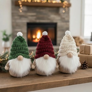 Może przedstawiać: Trzy ręcznie robione gnomy na szydełku z białymi brodami i drewnianymi nosami. Gnomy mają zielone, bordowe i kremowe czapki z białymi pomponami. Stoją na drewnianej powierzchni, idealne do dekoracji świątecznych.