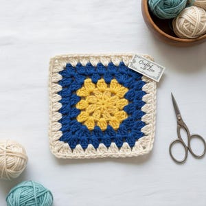 May include: A square, handmade crochet piece with a yellow center, surrounded by blue and cream-colored yarn. A tag that says "Crafted with Love" is attached. A pair of small scissors and balls of yarn are also in the image.
