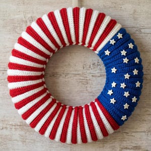May include: A circular wreath featuring a crocheted design with alternating red and white vertical stripes. One section of the wreath is solid blue and decorated with small, white, four-pointed star-shaped buttons arranged across the surface.