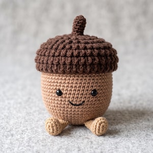 May include: A crocheted acorn toy featuring a dark brown textured cap and a light tan body. The acorn has two small black bead eyes and a stitched black smile, with two small tan feet extending from the bottom of the rounded body.