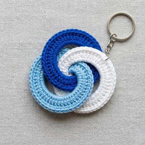 Peut inclure: Un porte-clés crocheté à la main avec trois anneaux entrelacés bleus et blancs. Les anneaux sont reliés à un porte-clés et une chaîne argentés. Le porte-clés est fait de fil doux et présente un aspect texturé.