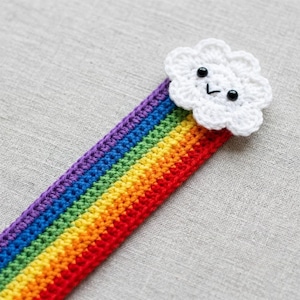Op de afbeelding: Een handgemaakte gehaakte bladwijzer met een regenboogontwerp en een lachende witte wolk aan de bovenkant. De regenboog loopt van rood naar oranje, geel, groen, blauw en paars. De bladwijzer is gemaakt van garen en ligt op een grijze ondergrond.