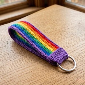 May include: A crocheted wristlet keychain featuring a vibrant rainbow pattern with horizontal stripes in purple, blue, green, yellow, orange, and red. The fabric loop is secured with a purple crocheted tab and attached to a circular silver metal ring.