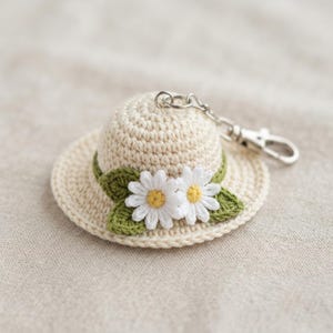 May include: A cream-colored crocheted hat keychain featuring two white daisy flowers with yellow centers and two green crocheted leaves attached to the base of the crown. The hat is connected to a silver-toned metal lobster clasp and ring.