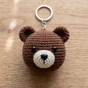 May include: A handmade brown crochet bear head keychain with a cream-colored muzzle and black eyes. The bear has a silver metal keyring attached to the top of its head. The keychain is a cute and cuddly accessory.