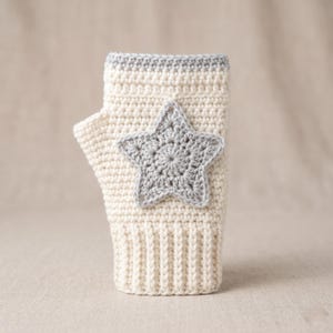Könnte beinhalten: Cremefarbener Fingerhandschuh aus Häkelarbeit mit einem grauen Stern-Applikation. Der Bund und der obere Rand sind mit grauem Garn verziert. Der Handschuh soll die Hände warm halten, während die Finger frei bleiben.