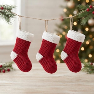 May include: Three small, handmade crochet Christmas stockings hanging from a thin twine string. Each stocking features a deep red body, a white cuff at the top, and a white heel section, all crafted with a consistent, textured yarn stitch pattern.