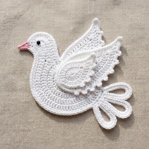 Può includere: Un'applicazione a forma di colomba lavorata all'uncinetto in bianco, con un piccolo becco rosa e un occhio a perla nera. L'uccello è raffigurato di profilo con le ali spiegate verso l'alto e una coda composta da tre anelli all'uncinetto.