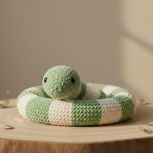 Puede incluir: Un juguete de serpiente de ganchillo en tonos verdes y blancos, enrollado sobre una superficie de madera. La cabeza de la serpiente es verde liso, con pequeños ojos negros. El cuerpo presenta un patrón de rayas. Un juguete suave hecho a mano.