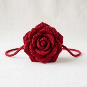 Puede incluir: Una rosa roja de ganchillo hecha a mano con un diseño detallado de pétalos. La rosa es tridimensional y presenta un asa de cordón rojo a juego, perfecta para un bolso o un elemento decorativo. La rosa está hecha de hilo suave.