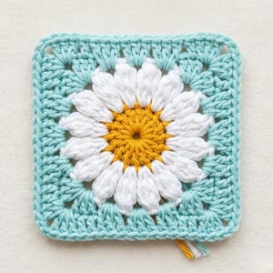 Puede incluir: Una pieza cuadrada de ganchillo con un diseño de margarita. El centro de la margarita es amarillo, rodeado de pétalos blancos. El fondo es azul claro, con un patrón de puntadas texturizadas. Una pequeña borla de hilo cuelga de la parte inferior.