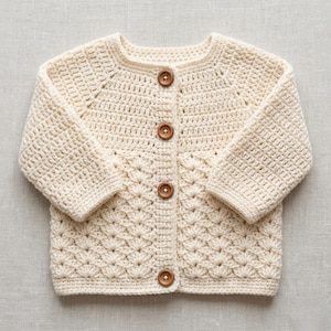 May include: A cream-colored long-sleeve cardigan made of crocheted yarn. The garment features a round neckline, long sleeves, and a front button closure with four round, light brown wooden buttons. The lower body has a textured shell stitch.
