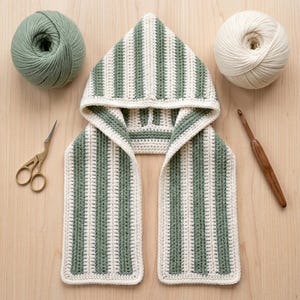 May include: A hooded scarf featuring a vertical striped pattern in sage green and cream yarn. The crochet garment includes a pointed hood and two long rectangular panels. Beside the scarf are two yarn balls, a pair of gold scissors, and a wooden hook.