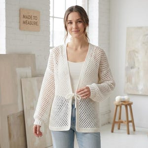 Op de afbeelding: Een witte, open geweven cardigan met lange mouwen en een striksluiting. De cardigan wordt gedragen over een witte tanktop en gecombineerd met een blauwe jeans. Een houten bord met de tekst "MADE TO MEASURE" is zichtbaar op de achtergrond.