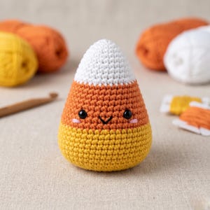 May include: A handmade crochet candy corn with a smiling face. The amigurumi features a white top, orange middle, and yellow bottom. Black safety eyes and pink cheeks complete the design. Yarn and a crochet hook are in the background.