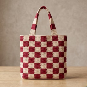 May include: A handmade crochet tote bag with a red and cream checkerboard pattern. The bag has a rectangular shape with a matching handle. The bag is sitting on a wooden surface.