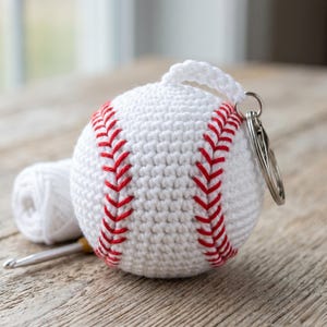 Puede incluir: Un llavero de béisbol redondo tejido a ganchillo en color blanco con detalles de costuras rojas. La pelota de béisbol está unida a una anilla de metal plateado. Un pequeño ovillo de lana blanca y un ganchillo de metal están junto a la pelota.