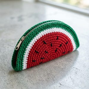 May include: A crocheted watermelon-shaped pouch with a silver zipper. The pouch features a red center with black seed details, bordered by a white stripe and a green rind. The handmade accessory is designed to resemble a slice of watermelon.