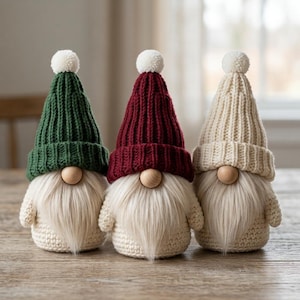 Könnte beinhalten: Drei Wichtelfiguren mit langen Kunstfellbärten und runden Holznasen. Jeder Wichtel trägt eine gerippte Strickmütze mit einem weißen Bommel. Die Mützen sind dunkelgrün, tiefrot und cremefarben, während die Körper aus cremefarbenem Häkelgarn bestehen.